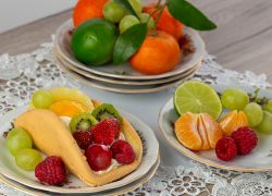 Fotografia kulinarna przedstawia ciasto owocowe języczek z truskawkami i malinami. W tle pomarańcze i winogrona